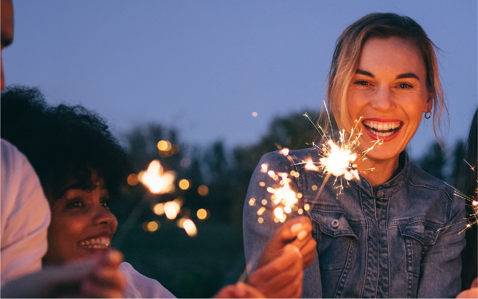 Young, white blonde woman, without glasses, holding sparkler with nearby friends